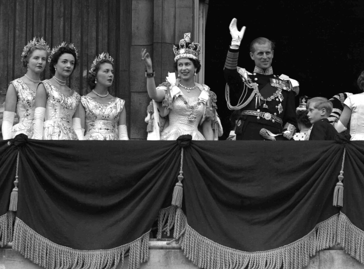 La reina Isabel II de Gran Bretaña y el príncipe Felipe saludan desde un balcón mientras tres mujeres miran cerca.