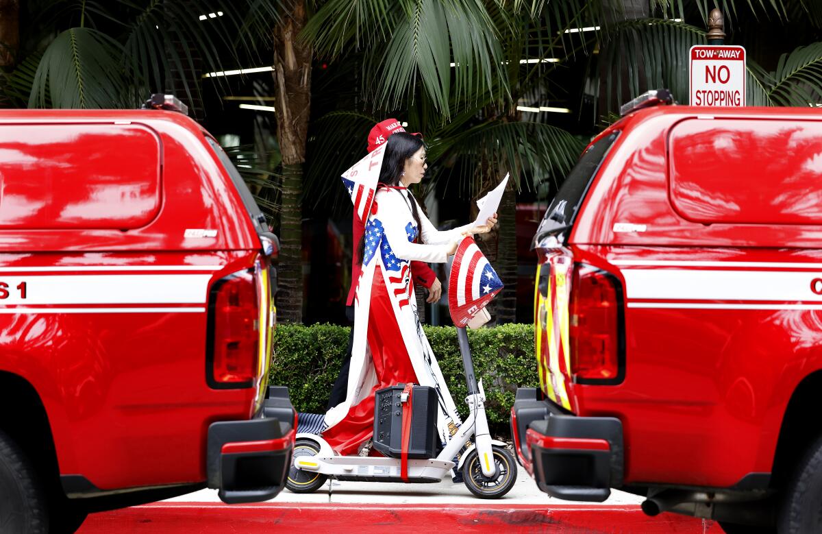 Una mujer vestida de rojo, blanco y azul vista en el espacio entre dos vehículos rojos mientras pasa con un altavoz atado a un scooter.