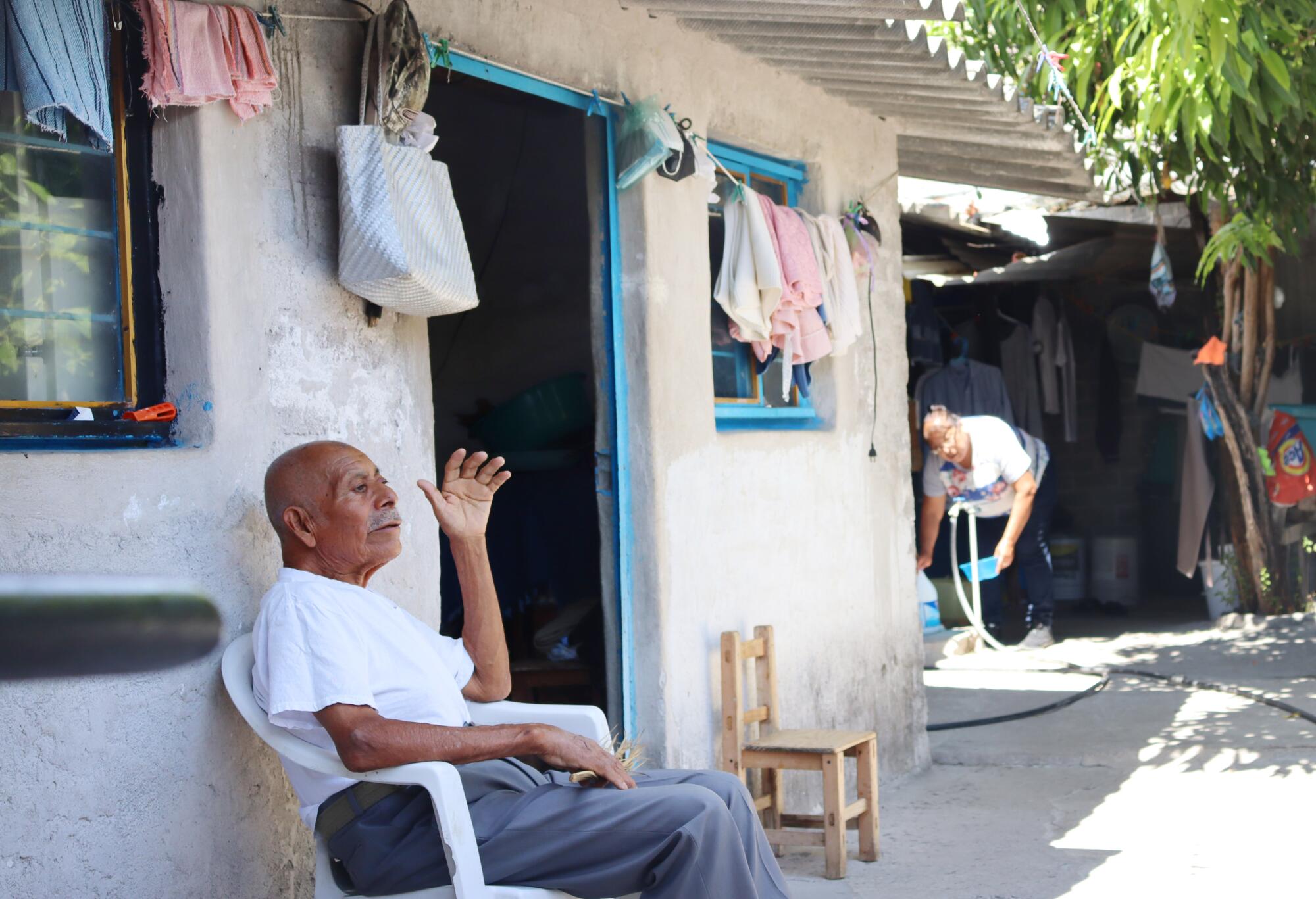 An older man with mustache, in a white T-shirt and gray pants, sits in a chair outside his home 